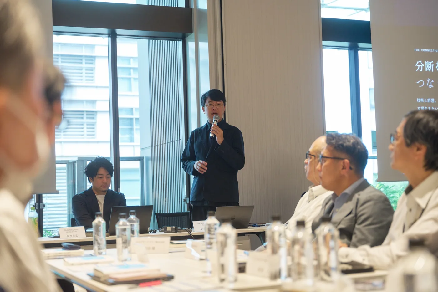 企業の役員が囲むテーブルの前に立って話す小澤健祐