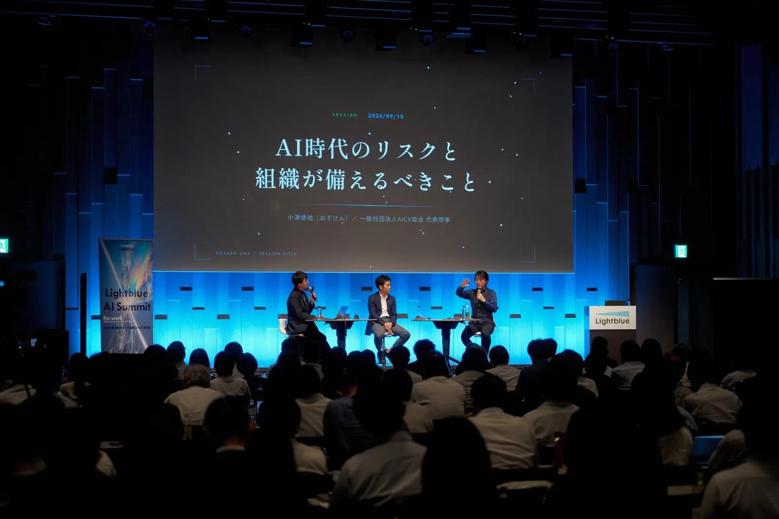 カンファレンスのパネルセッションで、登壇者2人と並んでマイクを持つ小澤健祐。スクリーンに「AI時代のリスクと組織が備えるべきこと」